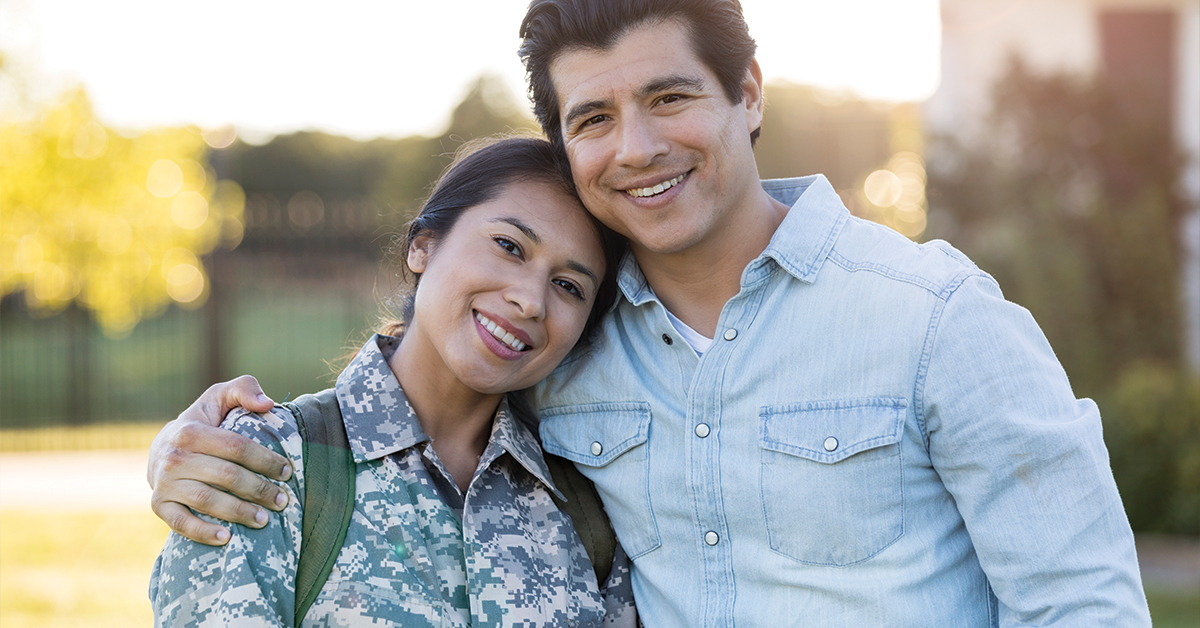 A mild adult couple stand in their front yard and embrace as they smile for the camera. The wife is in military camo and has just returned from a tour.