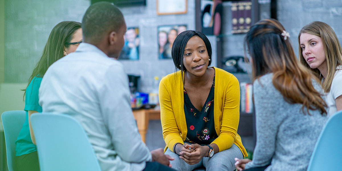 A diverse group of adults sit in a circle in their chairs and listen to and sympathize with each other with serious expressions.