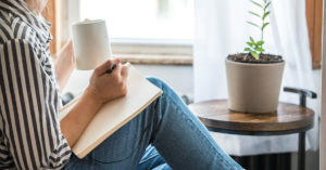 A young woman takes a break to do something analog like writing in her journal and drinking tea. This is a healthy practice for those who experience anxiety.