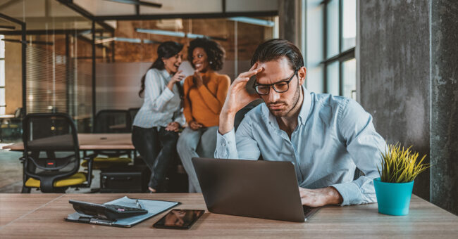Addressing Harassment Lonely and sad businessman at office of company. Photo of Sad businessman with female team in the background. Male office manager unable to cope with too many tasks from his boss and supervisor, suffering stress, pressing her head with desperate expression.