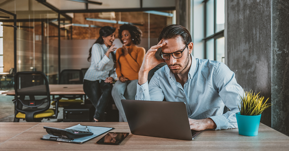 Lonely and sad businessman at office of company. Photo of Sad businessman with female team in the background. Male office manager unable to cope with too many tasks from his boss and supervisor, suffering stress, pressing her head with desperate expression.