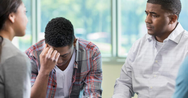 Blog Featured Image – Coping with the Loss of a Colleague Young African American man has his head in his hands during support group meeting. His friends are sitting around him.