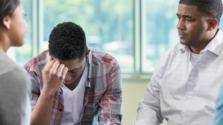 Blog Featured Image – Coping with the Loss of a Colleague Young African American man has his head in his hands during support group meeting. His friends are sitting around him.