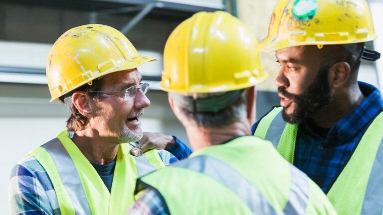Blog Featured Image – Handling a Confrontation A group of three multi-ethnic men wearing hard hats and safety vests having a meeting. They are serious expressions on their faces, perhaps having a disagreement. The focus is on the two men in the background.