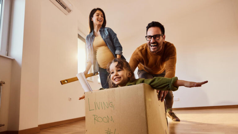Blog Featured Image – Make Your Next Move a Success Happy expecting family having fun after moving into a new home. Father is pushing their daughter in cardboard box.