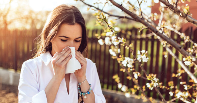 Blog Featured Image – Managing Allergy Symptoms Attractive young adult woman coughing and sneezing outdoors. Sick people allergy or virus influenca concept.