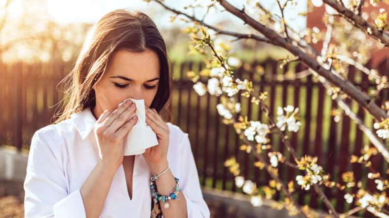 Blog Featured Image – Managing Allergy Symptoms Attractive young adult woman coughing and sneezing outdoors. Sick people allergy or virus influenca concept.