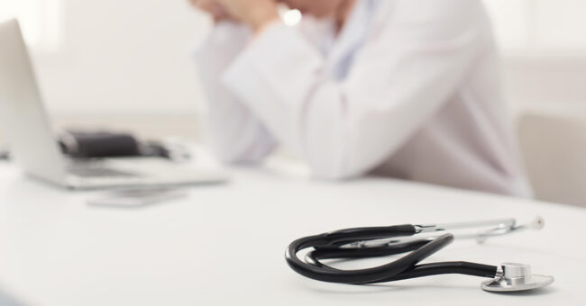 Blog Featured Image – Mental Health at Work Unrecognizable tired doctor sitting at workplace. Overworked physician with headache resting in modern office, medicine concept, copy space