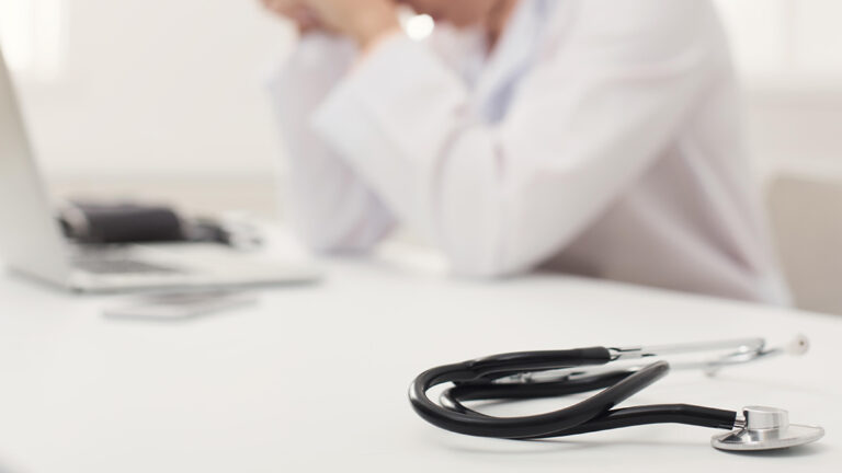 Blog Featured Image – Mental Health at Work Unrecognizable tired doctor sitting at workplace. Overworked physician with headache resting in modern office, medicine concept, copy space