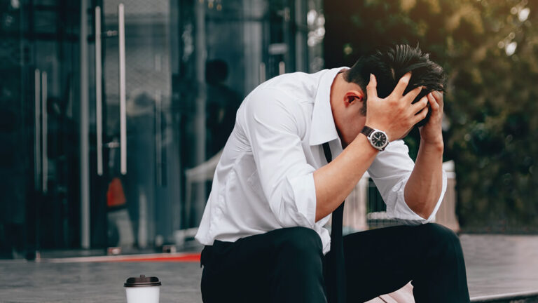 Coping with Survivor’s Guilt Asian young businessman stress sitting in front office with his hands covering his head against.