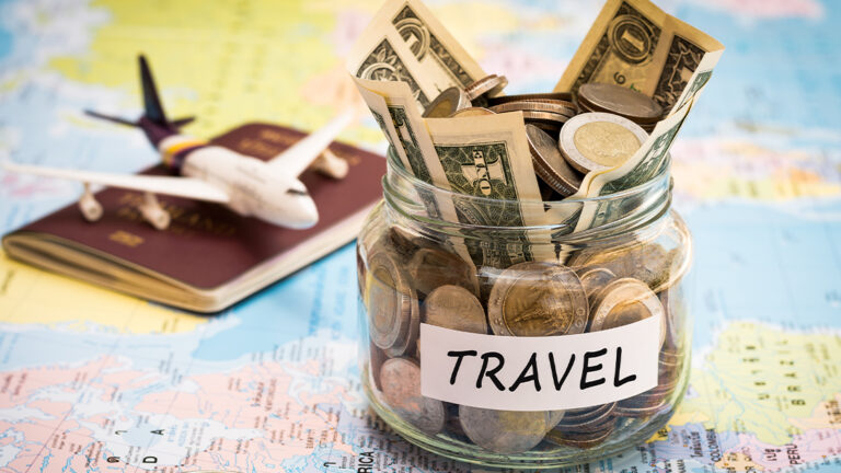 Blog Featured Image_Saving for Vacation Travel budget concept. Travel money savings in a glass jar with compass, passport and aircraft toy on world map
