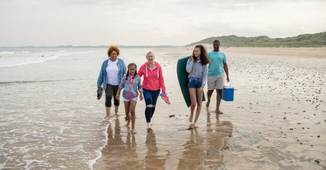 Decreasing Vacation Costs A multi-generation family walking side by side on Beadnell beach, North East England, in the edge of the sea. They are carrying a picnic box and paddle board between them.
