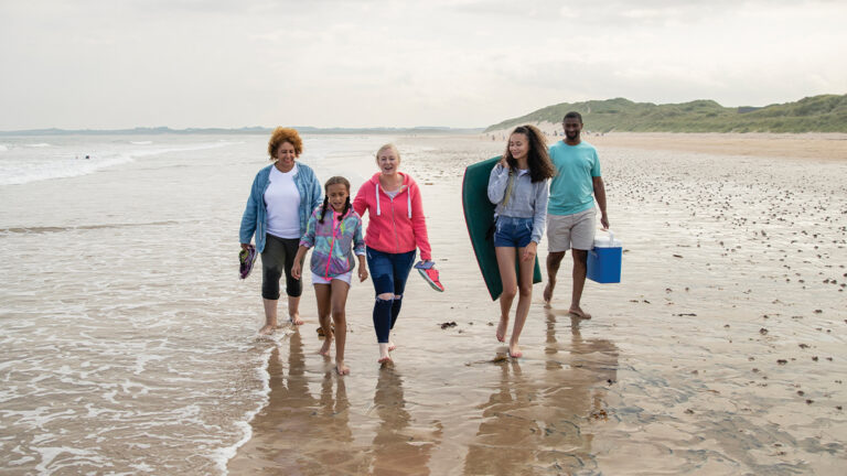 Decreasing Vacation Costs A multi-generation family walking side by side on Beadnell beach, North East England, in the edge of the sea. They are carrying a picnic box and paddle board between them.