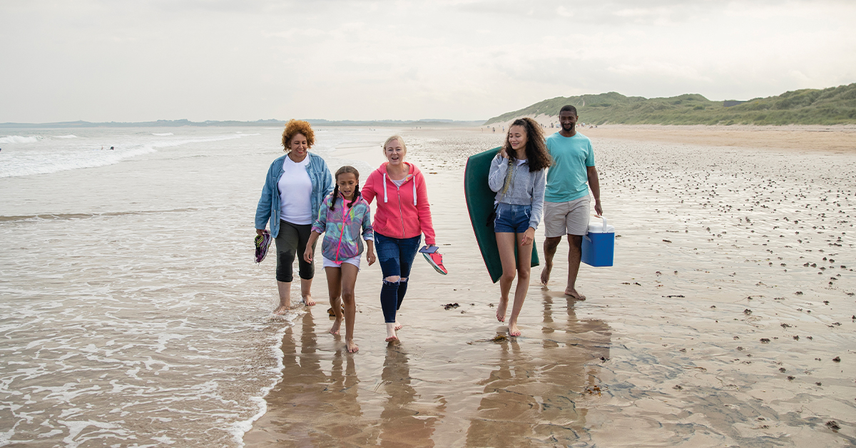 A multi-generation family walking side by side on Beadnell beach, North East England, in the edge of the sea. They are carrying a picnic box and paddle board between them.