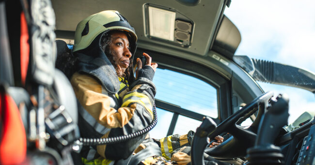 Featured Image – Episode 29- First Responder Mental Health Pt 2 Side view of a Black female airport firefighter sitting in a fire truck using a CB station while driving towards an emergency location at the airport runway.