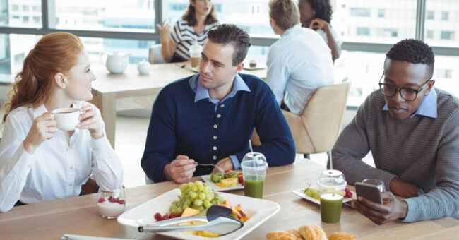 Featured Image – Respectful Workplace Behavior During an Election colleagues from mixed race backgrounds indulging in lunch in a contemporary office space. Seated at a table, engaging in conversation about the election and appreciating shared moments.