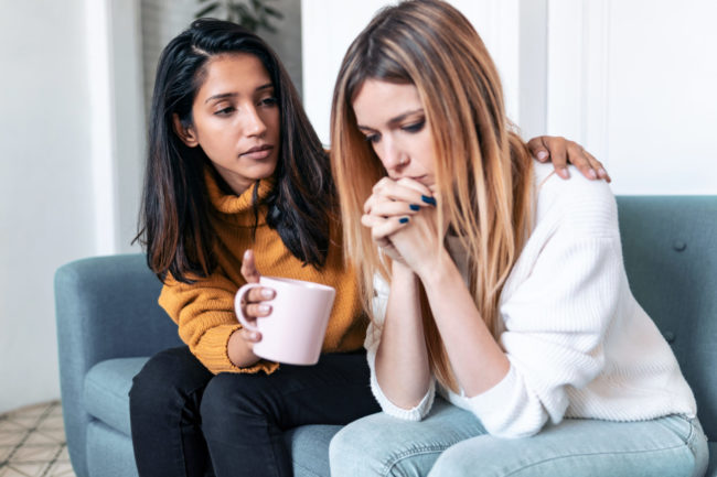 Pretty young woman supporting and comforting her sad friend while sitting on the sofa at home. one woman consoling another