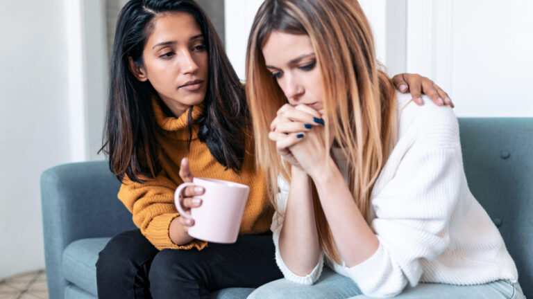 Pretty young woman supporting and comforting her sad friend while sitting on the sofa at home. one woman consoling another