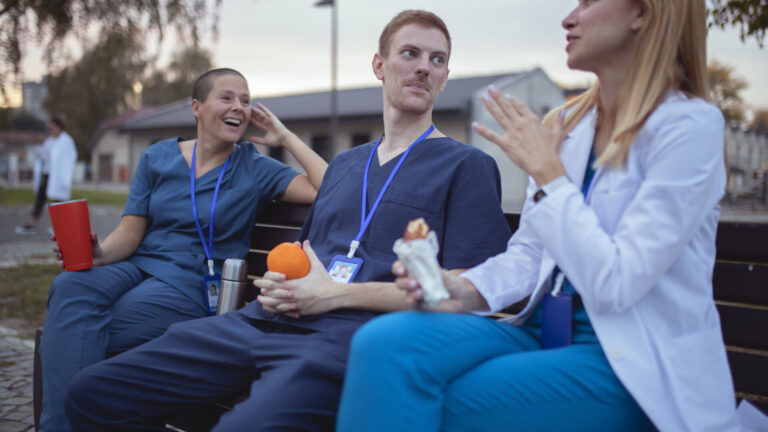 A doctor in a white lab coat talks to her colleagues in uniform, while they sit on a bench A small group of medical workers sits on a bench in the hospital yard and chats during a coffee break