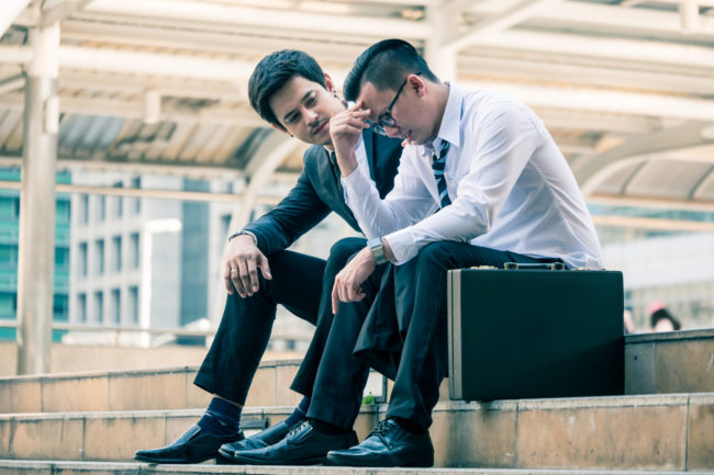 Frustrated Asian young business man sitting outdoor office after he had failed in new project, his colleagues tried to reassure his. one man consoling another