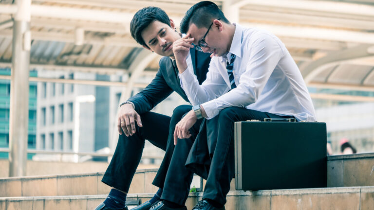 Frustrated Asian young business man sitting outdoor office after he had failed in new project, his colleagues tried to reassure his. one man consoling another