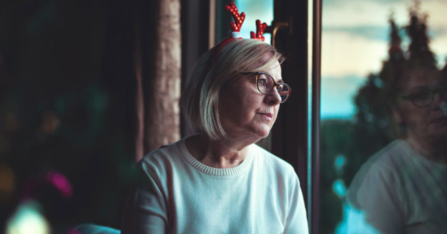 Loneliness-During-the-Holidays Senior woman who spent Christmas alone away from her family and friends due to the pandemic looks out the window sadly