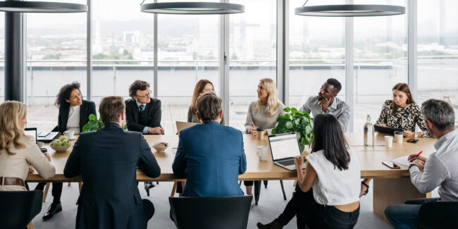 Management Consultation A photo of a business meeting in a modern office with large windows. A businessman is standing up while his colleagues are sitting down. They are smartly dressed. Horizontal daylight indoor photo.