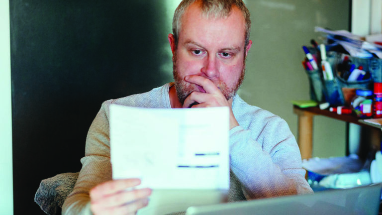 PayDown-Holiday-Debt Portrait of a mid adult man checking his energy bills at home. He has a worried expression and touches his face with his hand while looking at the bills. Focus on the man while the interior architecture of the house is defocused.