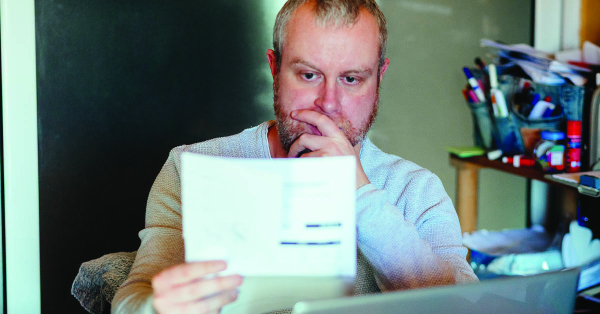 Portrait of a mid adult man checking his energy bills at home. He has a worried expression and touches his face with his hand while looking at the bills. Focus on the man while the interior architecture of the house is defocused.