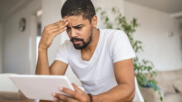 Soft-Skills-For-Management-Success Worried mixed ethnicity man sitting in a living room, contemplating with a digital tablet.