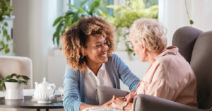 Female home caregiver talking with senior woman, sitting in living room and listening to her carefully.