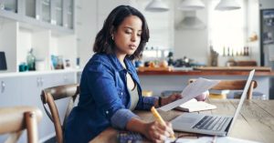 Shot of a young woman using a laptop and going through paperwork while working from home