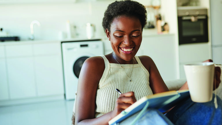 gratitude-journaling Shot of a young woman having tea and writing in a notebook on the sofa at home