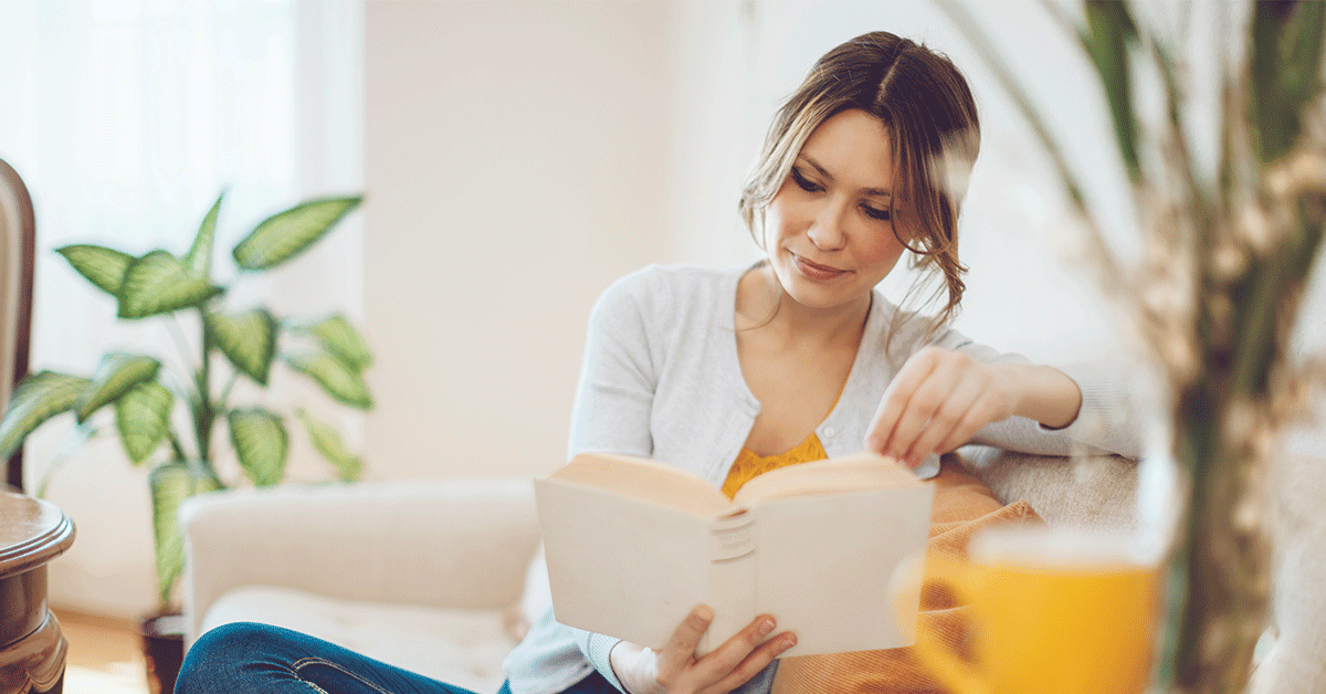 Content woman is reading a book on a white sofa at home. She is sitting comfortable and is wearing blue jeans and a grey sweater. The living room is cosy and elegant. Her hair and eyes are brown.