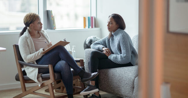 A Therapist meets with her female client in her office. The client is seated on a sofa with her arms across her body as she looks visibly nervous. The Therapist is seated in a chair in front of her as she talks about what to expect from the appointment and takes notes on her clipboard. A Therapist meets with her female client in her office. The client is seated on a sofa with her arms across her body as she looks visibly nervous. The Therapist is seated in a chair in front of her as she talks about what to expect from the appointment and takes notes on her clipboard.