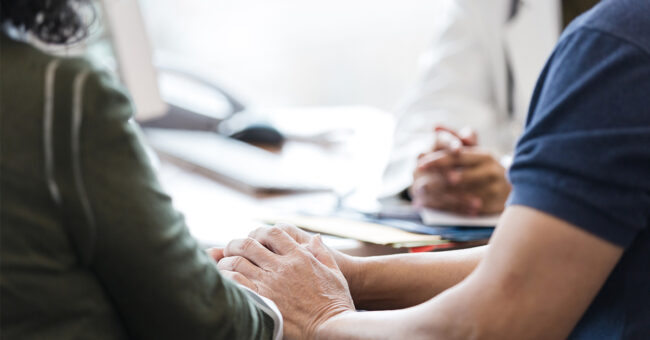 recognizable man holds his wife’s hand as she receives a difficult medical diagnosis while meeting with a doctor. recognizable man holds his wife's hand as she receives a difficult medical diagnosis while meeting with a doctor.