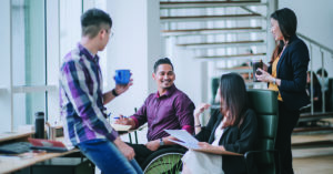 Indian white collar male worker in wheelchair having cheerful discussion leading conversation with colleague in creative office workstation beside window